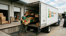 Logistics operators loading wooden crates of fresh regional produce into an INDYpendent Bytes box truck at a concrete loading dock hub.