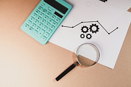 A top-down photograph on a tan surface showing a mint-green calculator, a magnifying glass with a black handle, and a white piece of paper featuring a printed line graph layered over an icon of three interlocking gears. >