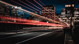 A long-exposure night photograph of an urban street flanked by illuminated city skyscrapers, showing vibrant red and white light trails from passing traffic cutting across the frame.